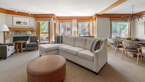 A modern living room with a sectional sofa, a round ottoman, a fireplace, and a dining area with wooden chairs and a table.