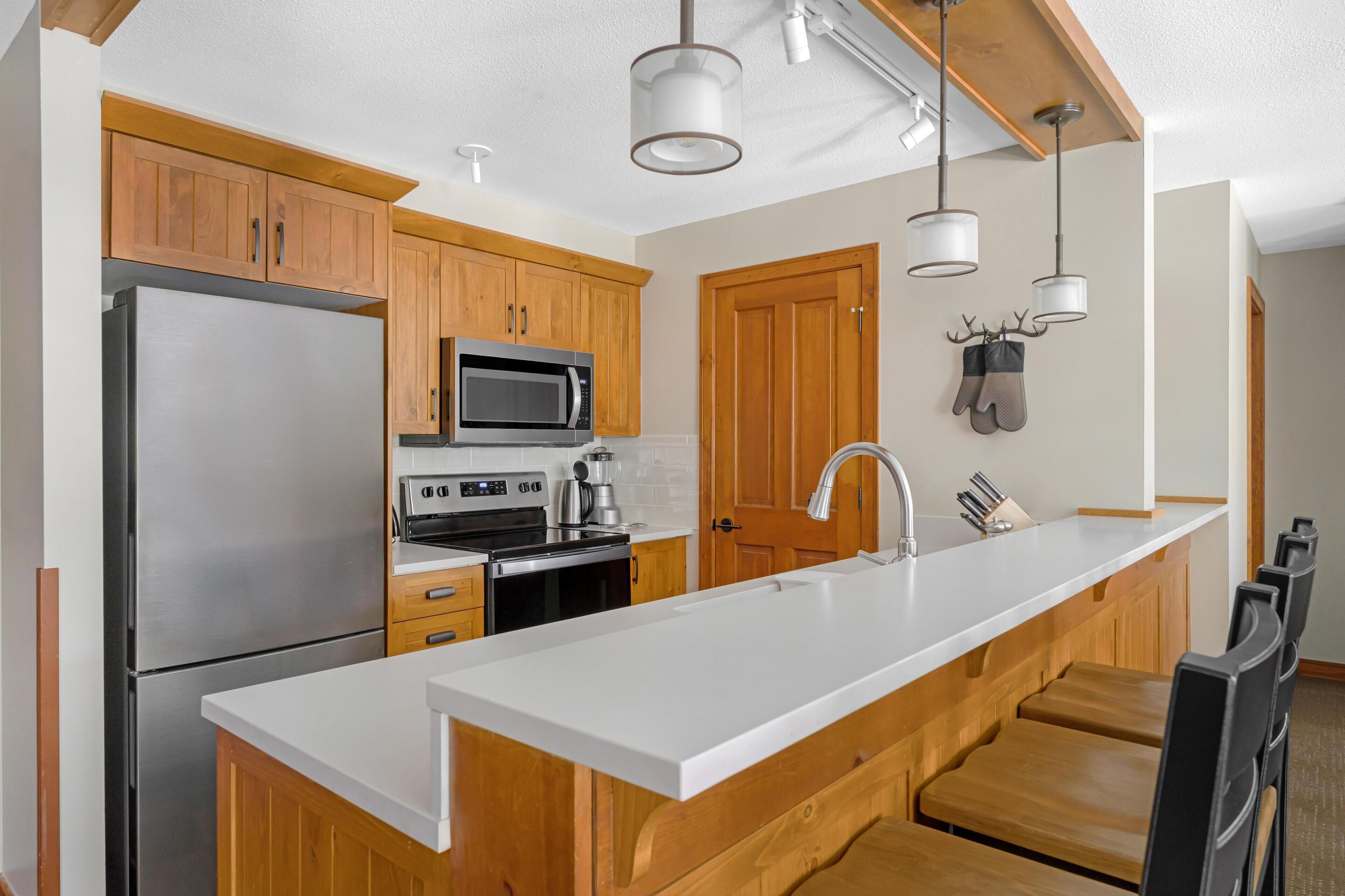 A modern kitchen with wooden cabinets, stainless steel appliances, a white countertop, and pendant lighting.