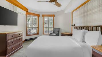 A hotel room with a large bed, a ceiling fan, a TV, two windows, a chair, and a bedside table.