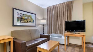 A hotel room with a brown leather sofa, a wooden coffee table, a TV on a wooden stand, a framed picture on the wall, and a window with curtains.