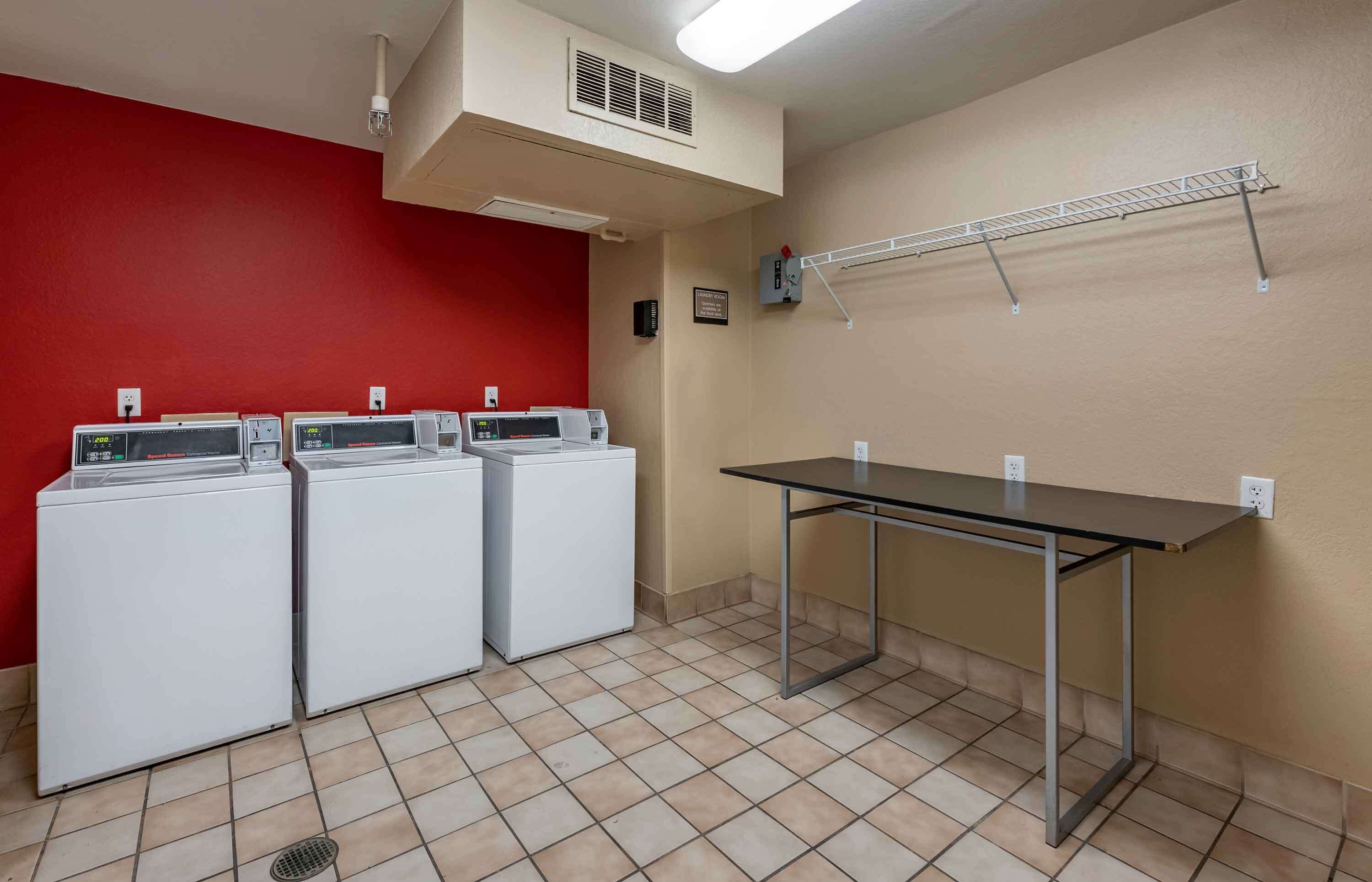 A laundry room with three white washers and dryers, a metal table, and a red wall.