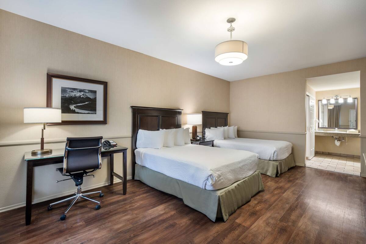 Habitación de hotel con dos camas, un escritorio con silla, una lámpara, un cuadro en la pared y un baño con espejo e iluminación.