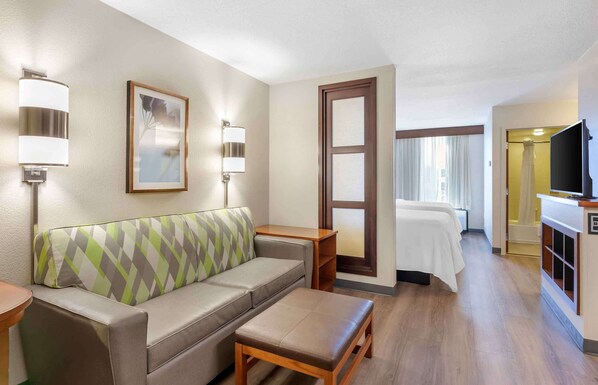 A hotel room with a sofa, a small table, a bench, a bed, a TV, and a bathroom with a shower.