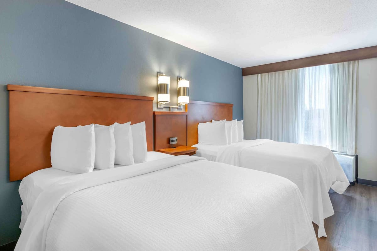 A hotel room with two beds, a wooden headboard, a nightstand with a lamp, and a window with curtains.