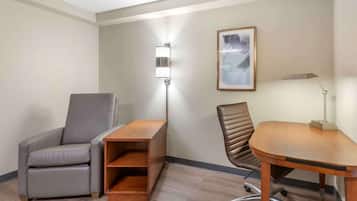 A hotel room with a grey leather armchair, a wooden desk, a standing lamp, and a framed abstract painting on the wall.