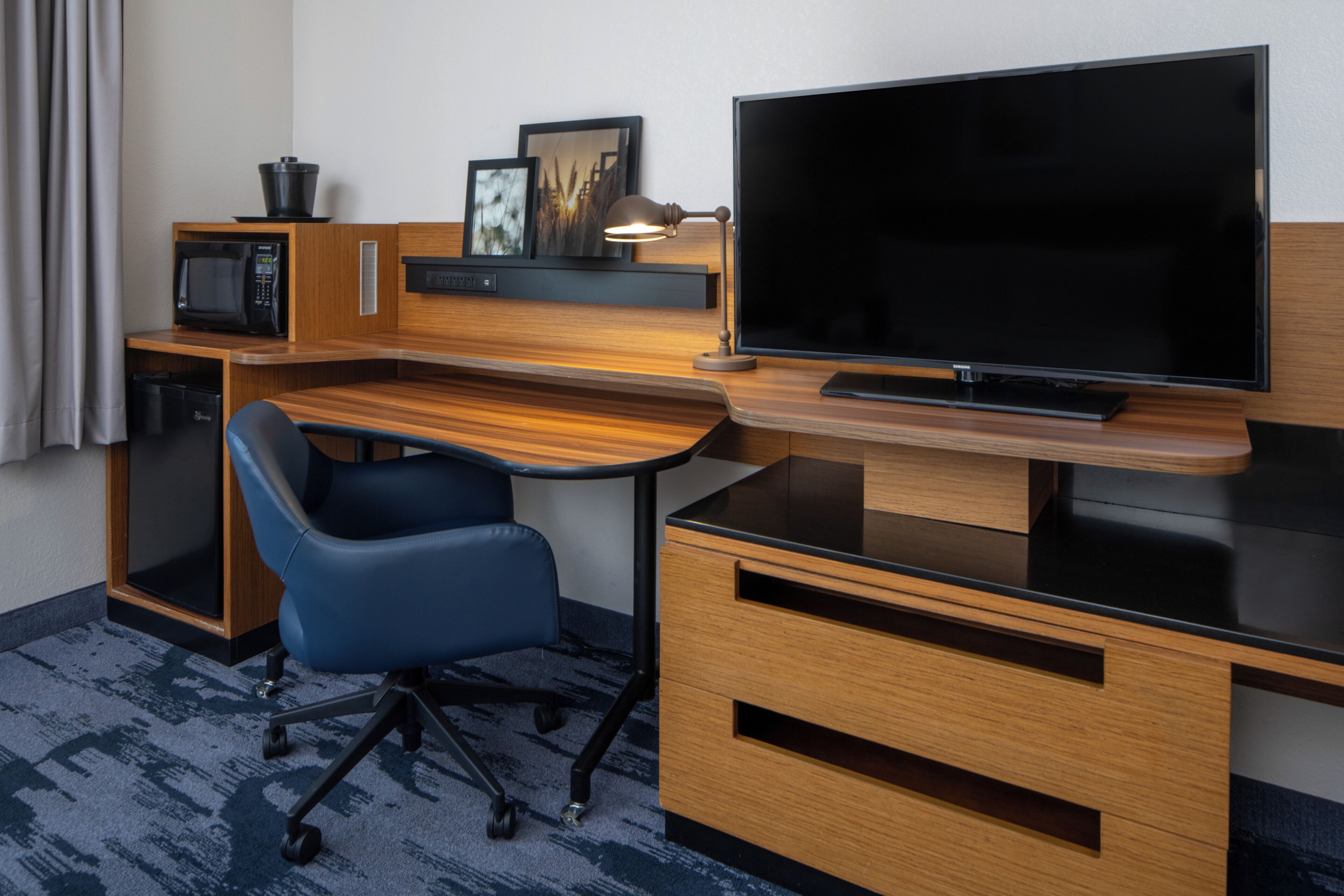 A hotel room with a wooden desk, a flat-screen TV, a microwave, a chair, and a small cabinet.