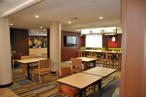 Un comedor de hotel con varias mesas y sillas, un televisor montado en la pared y un amplio ventanal con cortinas.