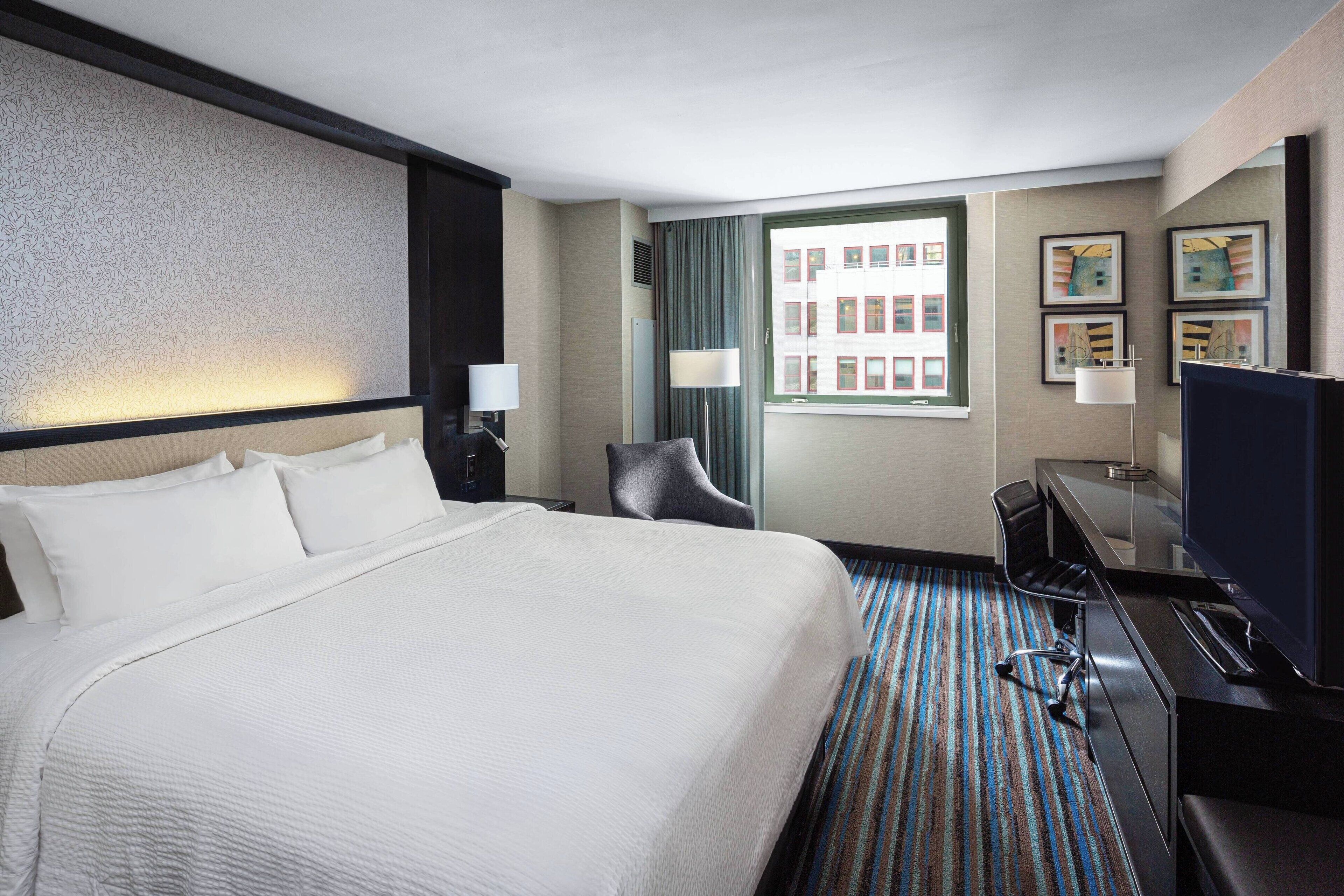 A hotel room with a large bed, a desk with a chair, a television, and a window with a view of buildings.