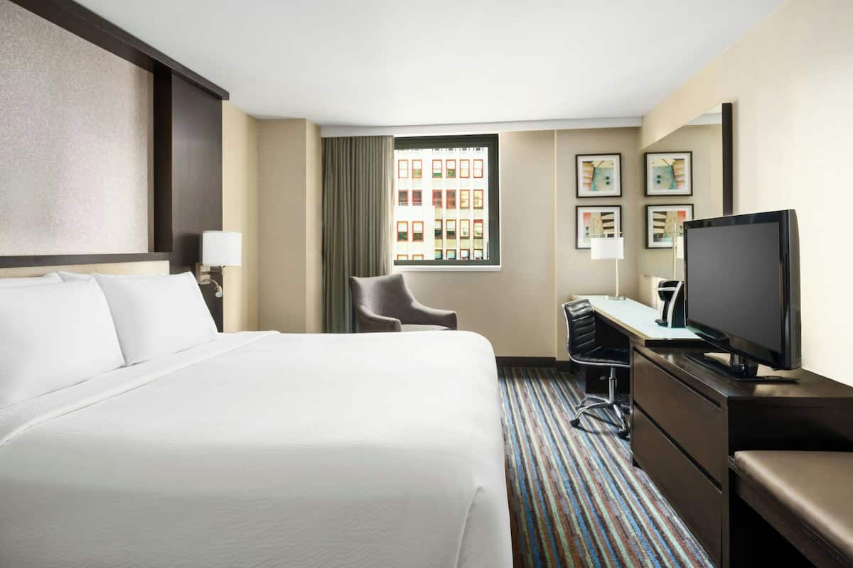 Habitación de hotel con una cama grande, un escritorio con televisor, una silla y una ventana con vistas a edificios.