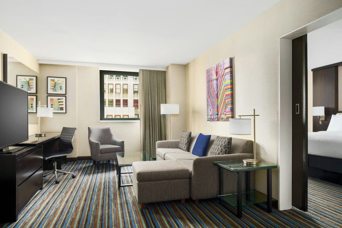 Habitación de hotel con sofá modular, escritorio con silla, televisor, mesa de centro de vidrio y cama.