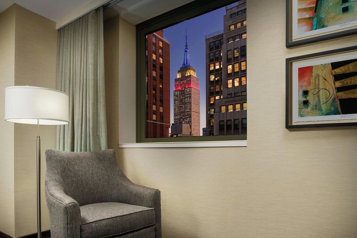 Habitación de hotel con vista al Empire State Building, un sillón gris, una lámpara de pie y dos cuadros en la pared.