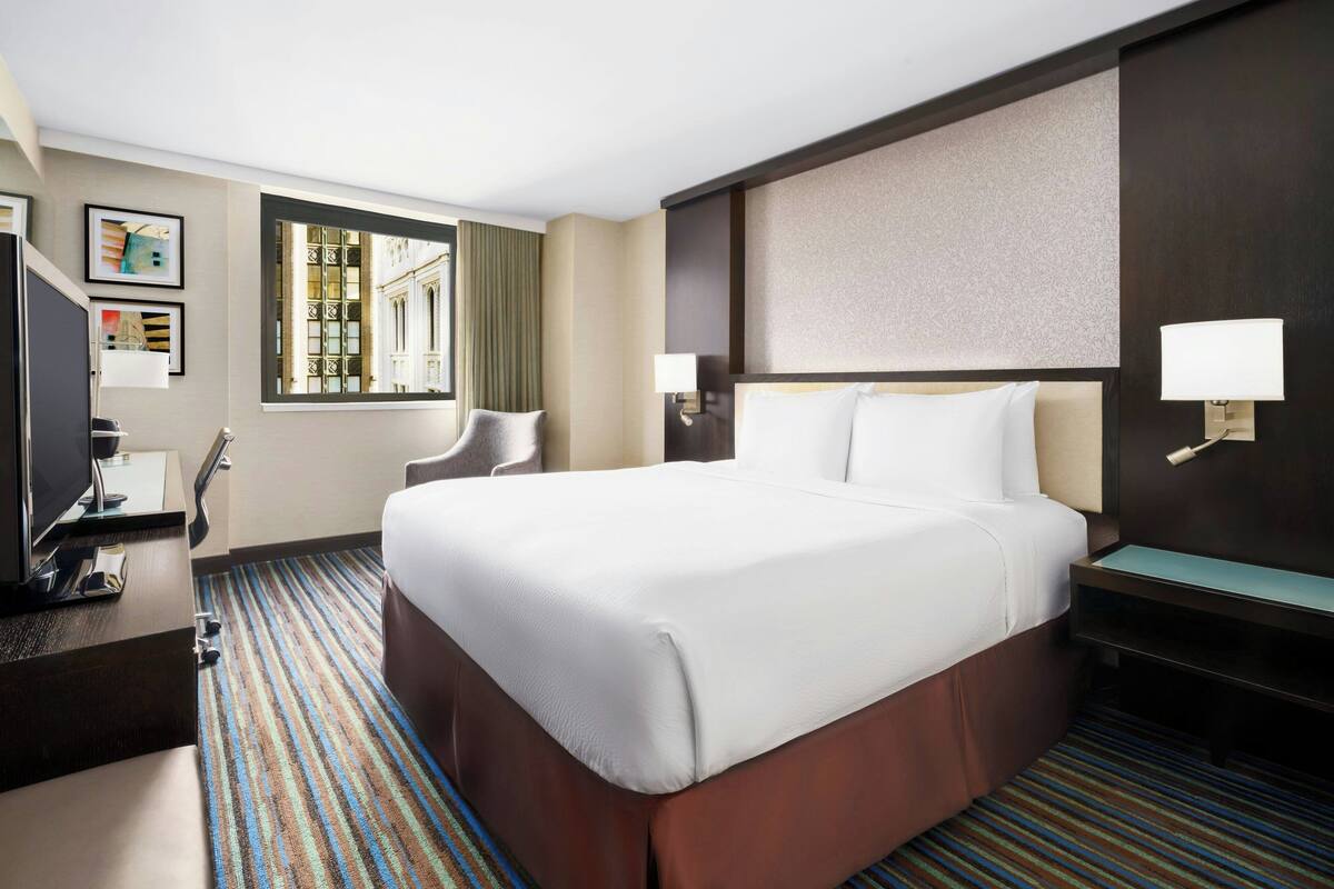 Habitación de hotel con una cama grande, un escritorio con computadora, una silla y una ventana con cortinas.