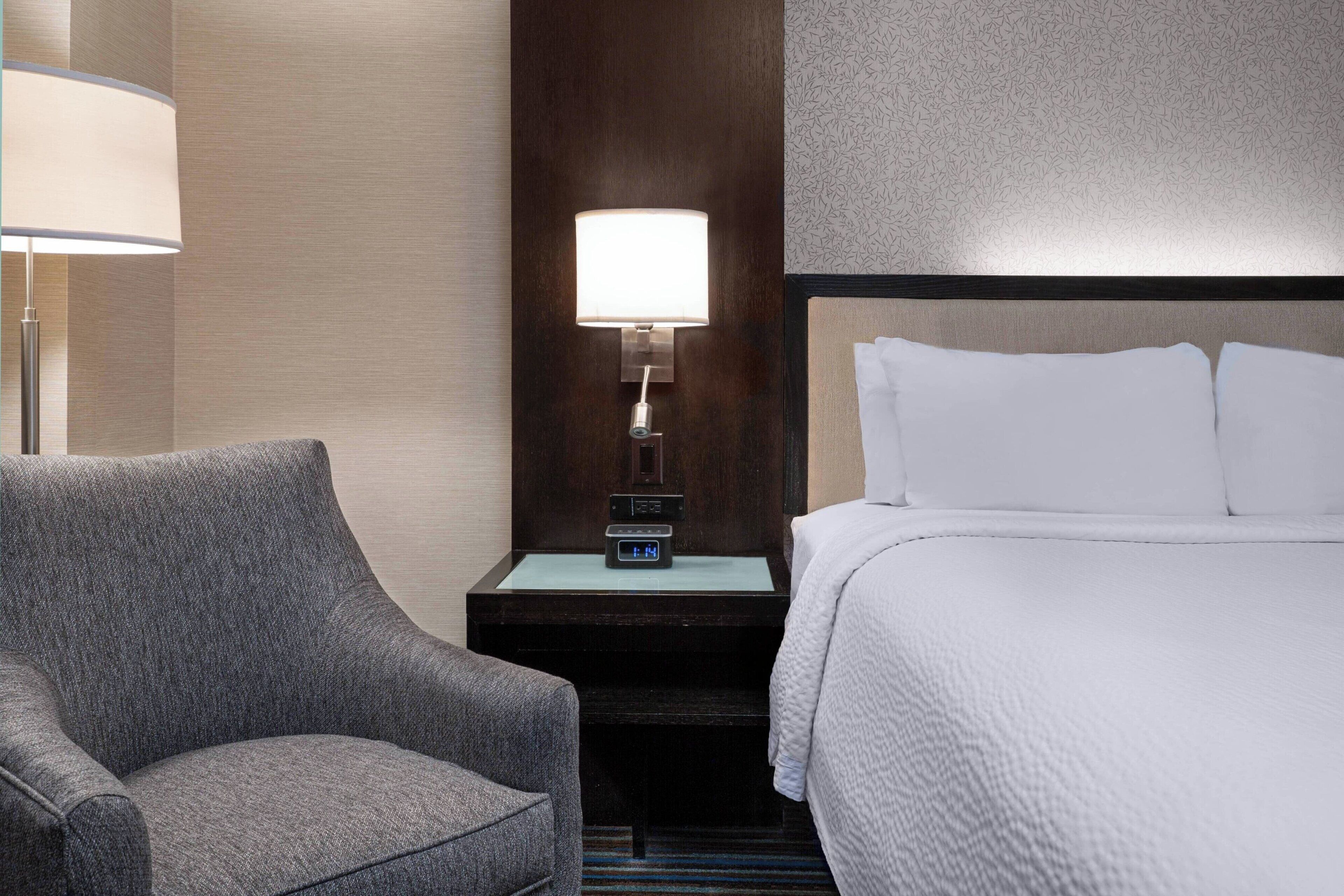 Una habitación de hotel con una cama, una mesita de noche con lámpara y un sillón gris.