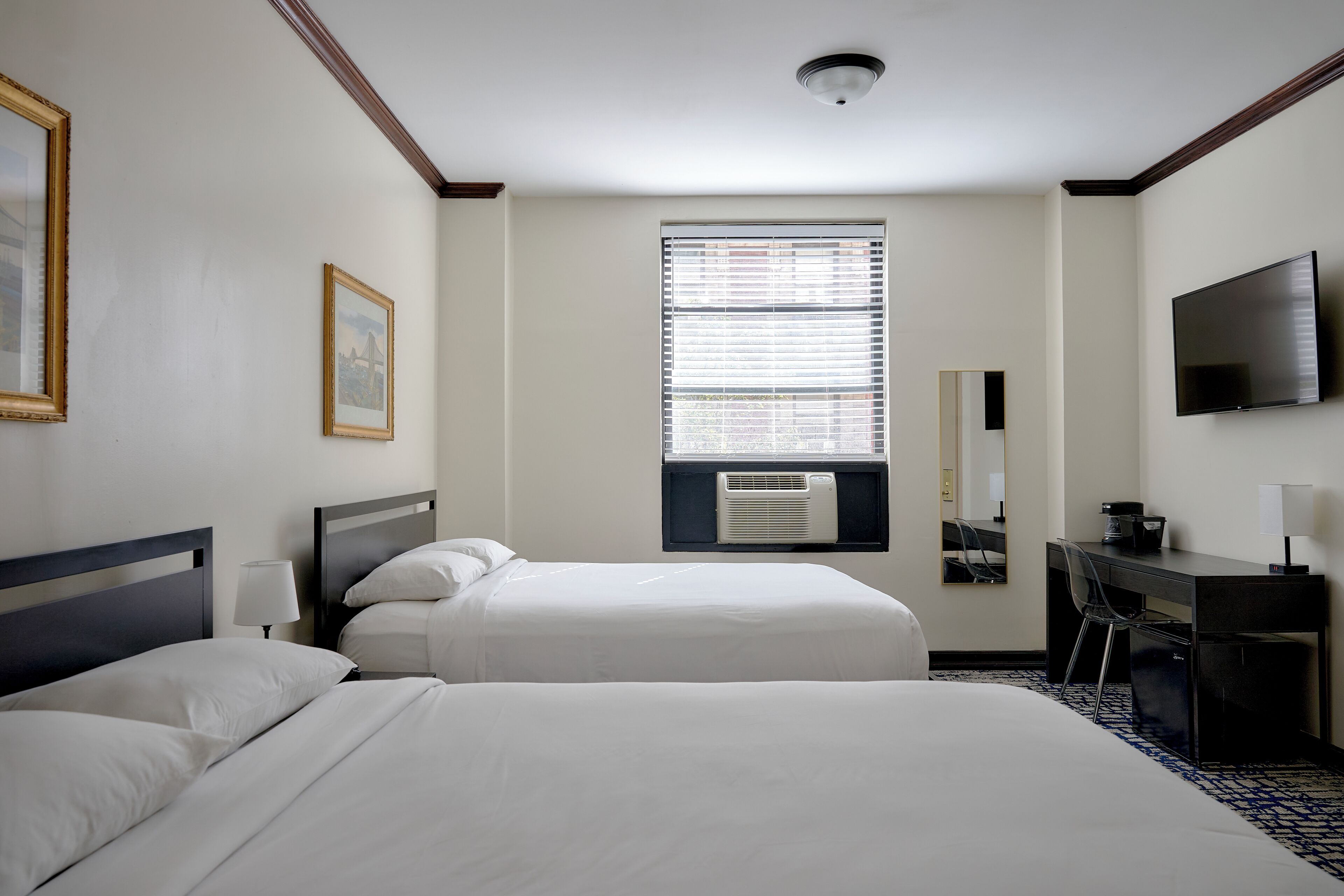Ein Hotelzimmer mit zwei Betten, einem Schreibtisch, einem Stuhl, einem Fernseher und einem Fenster mit Jalousien.