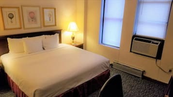 Una habitación de hotel con una cama grande, una mesita de noche con lámpara, una ventana con persianas y un equipo de aire acondicionado.