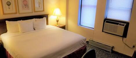 Quarto de hotel com uma cama grande, mesa de cabeceira com abajur, janela com persianas e um aparelho de ar condicionado.