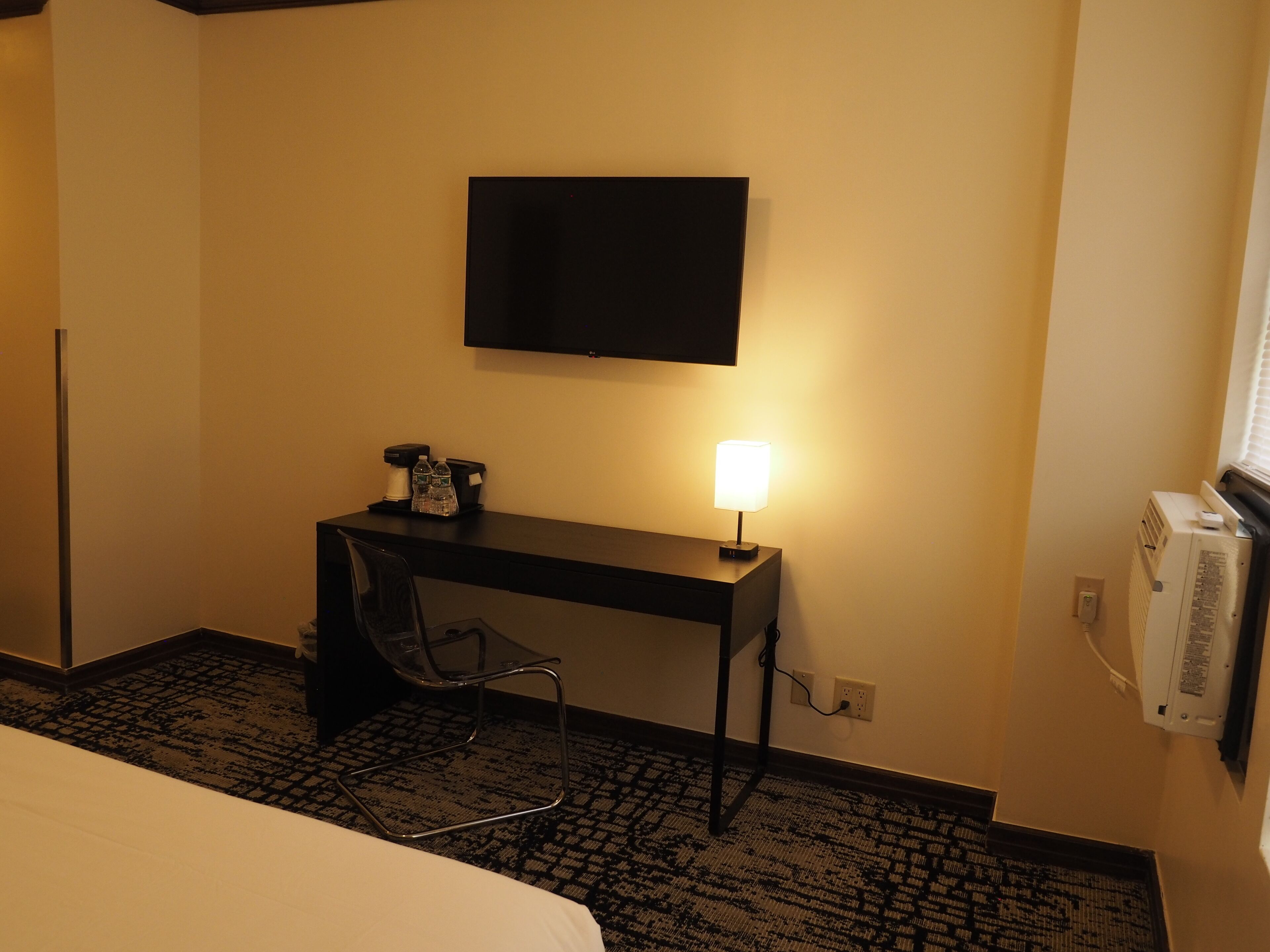 A hotel room with a bed, a desk with a lamp, a chair, a flat-screen TV mounted on the wall, and an air conditioning unit.
