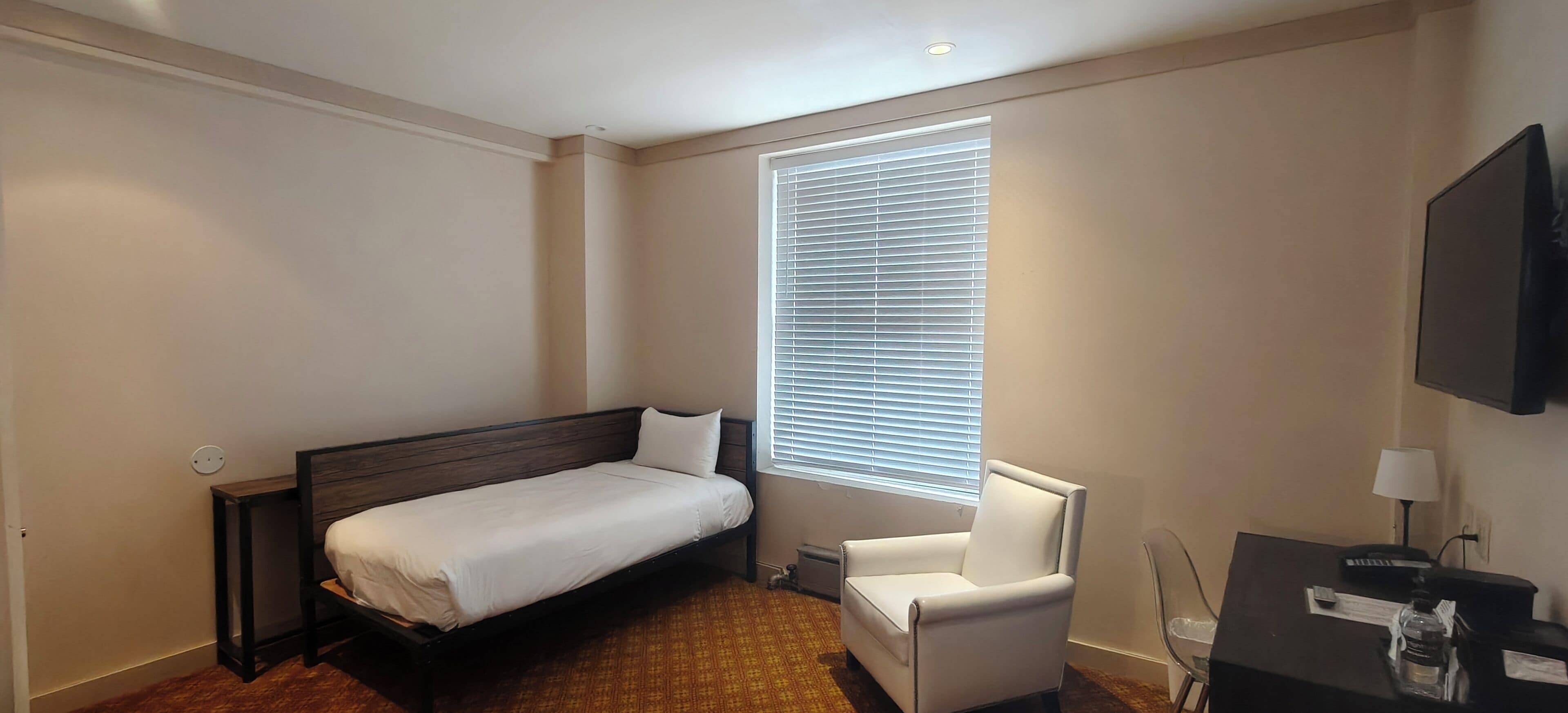 A hotel room with a single bed, a chair, a desk with a lamp, and a TV on the wall.