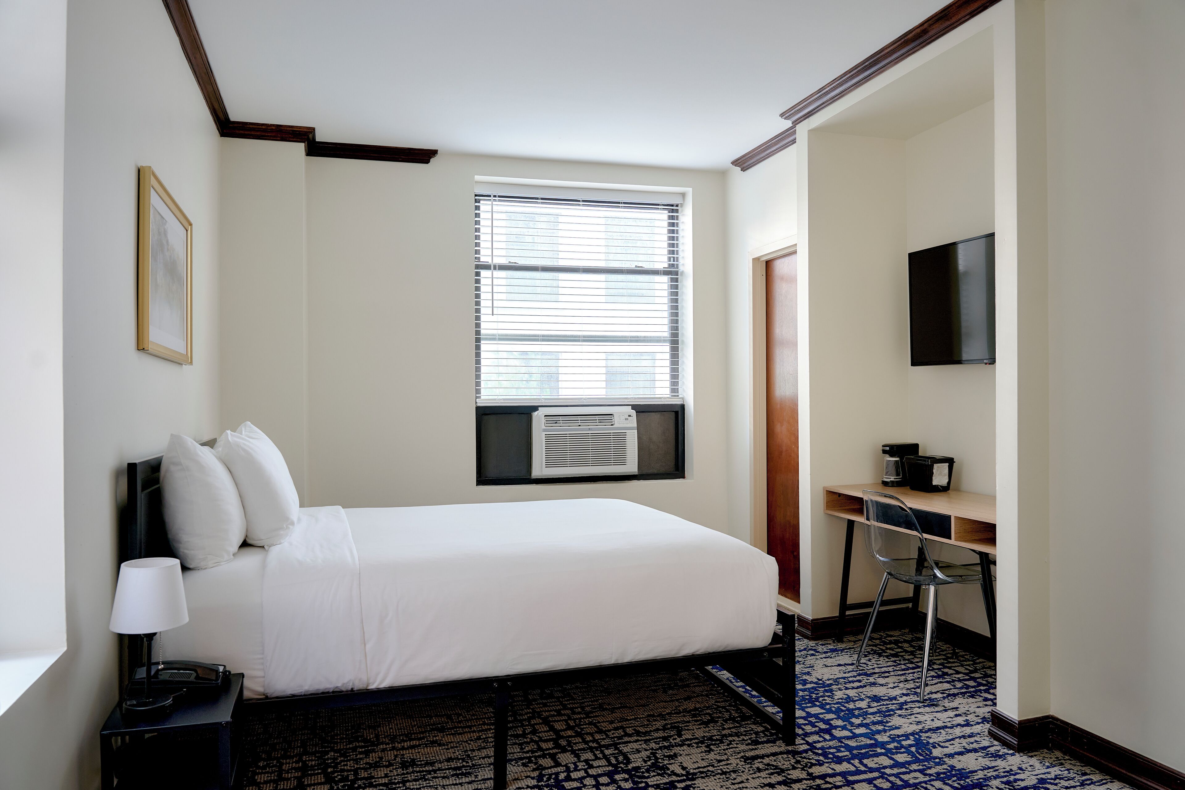 A hotel room with a bed, a desk, a chair, a TV, and an air conditioning unit.