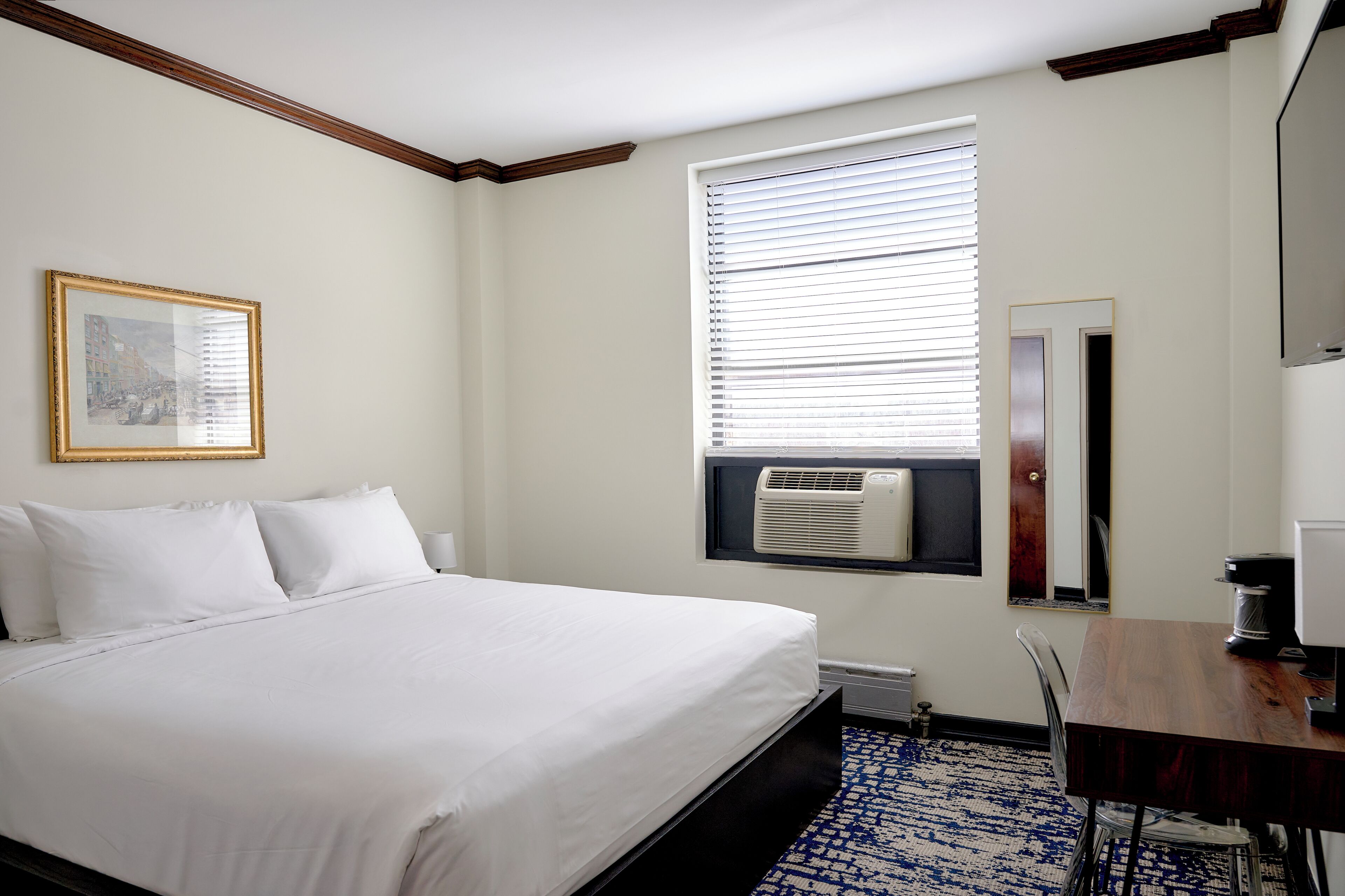 A hotel room with a bed, a window with blinds, a wall-mounted air conditioner, a framed picture, a door, and a small table with a coffee maker.