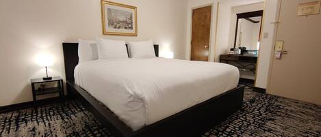 Un lit bien fait dans une chambre d’hôtel, avec une tête de lit sombre, une petite table de chevet avec une lampe, un tableau encadré au mur et une porte donnant sur une salle de bain.