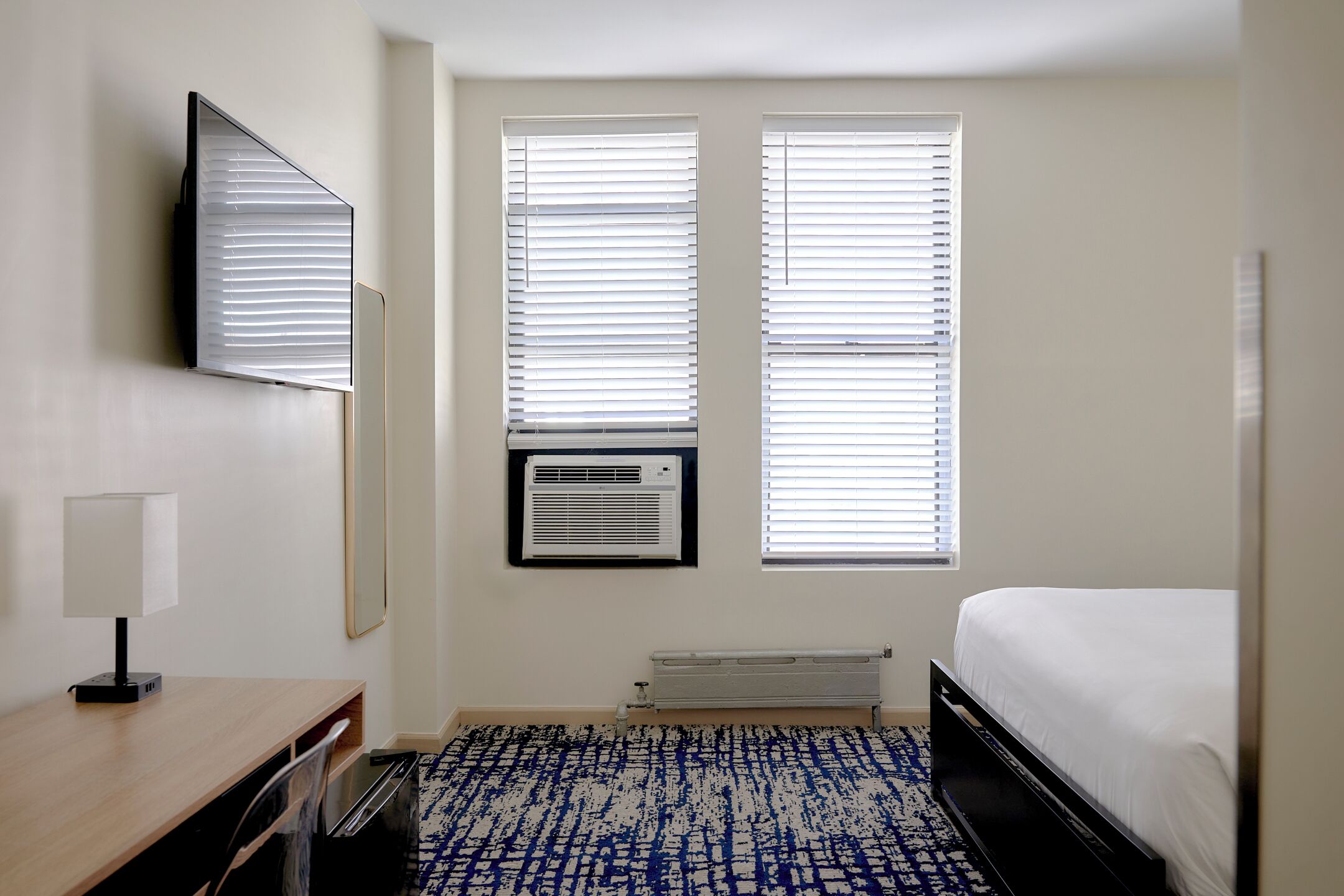 A hotel room with a bed, a desk with a lamp, two windows with blinds, an air conditioning unit, and a radiator.