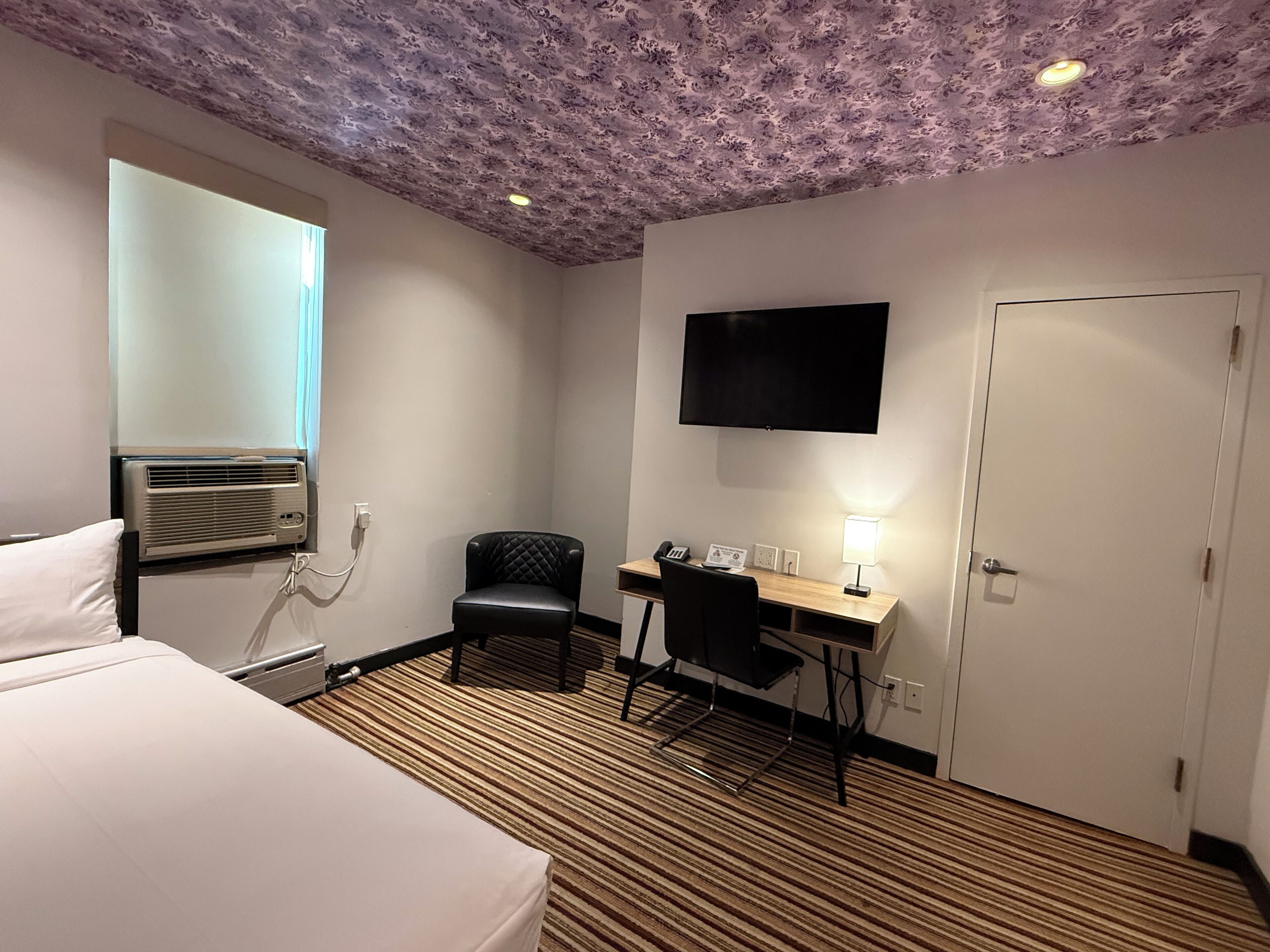 Habitación de hotel con cama, escritorio, silla, televisor y aire acondicionado.