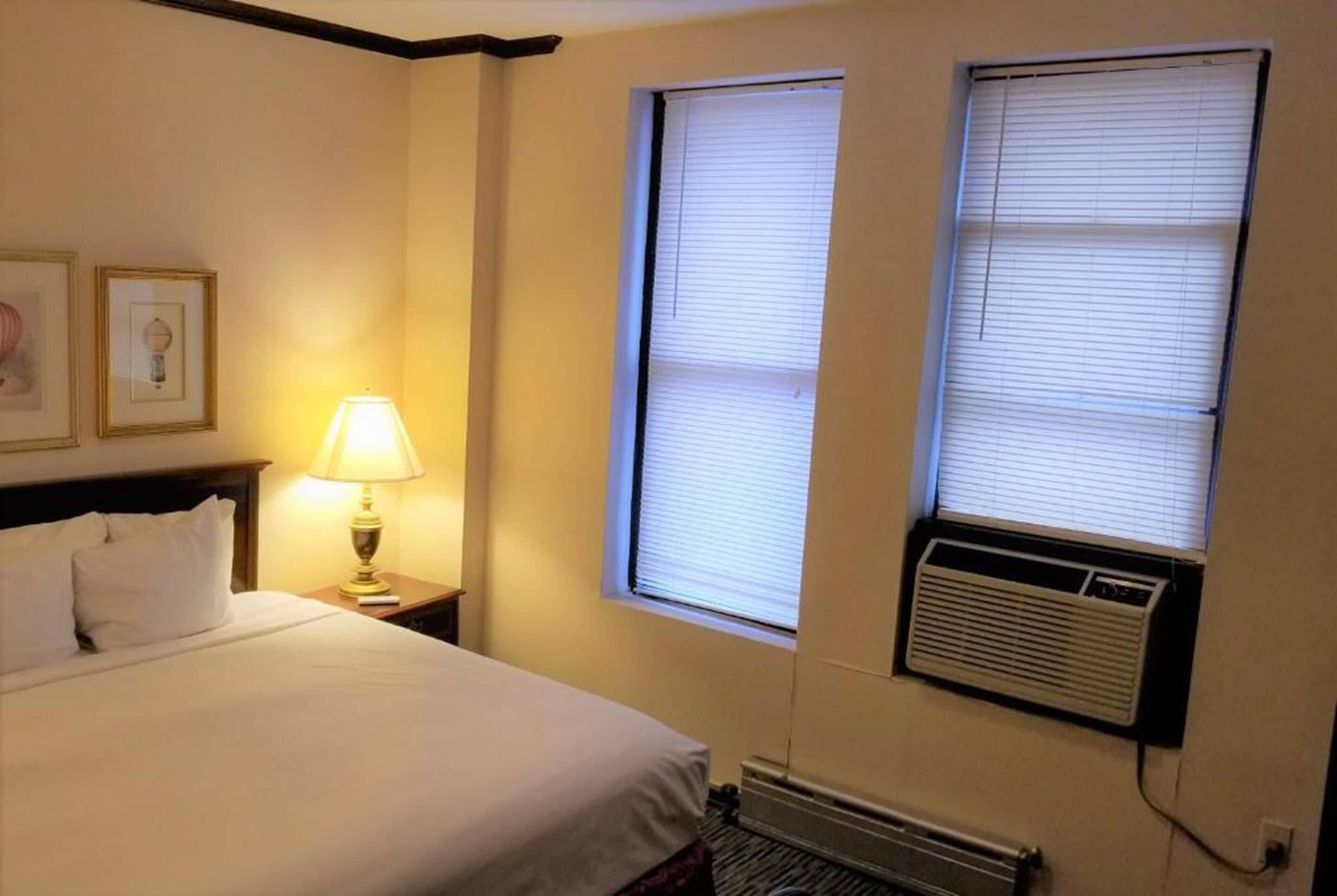 Habitación de hotel con cama, mesita de noche con lámpara, dos ventanas con persianas y un equipo de aire acondicionado.