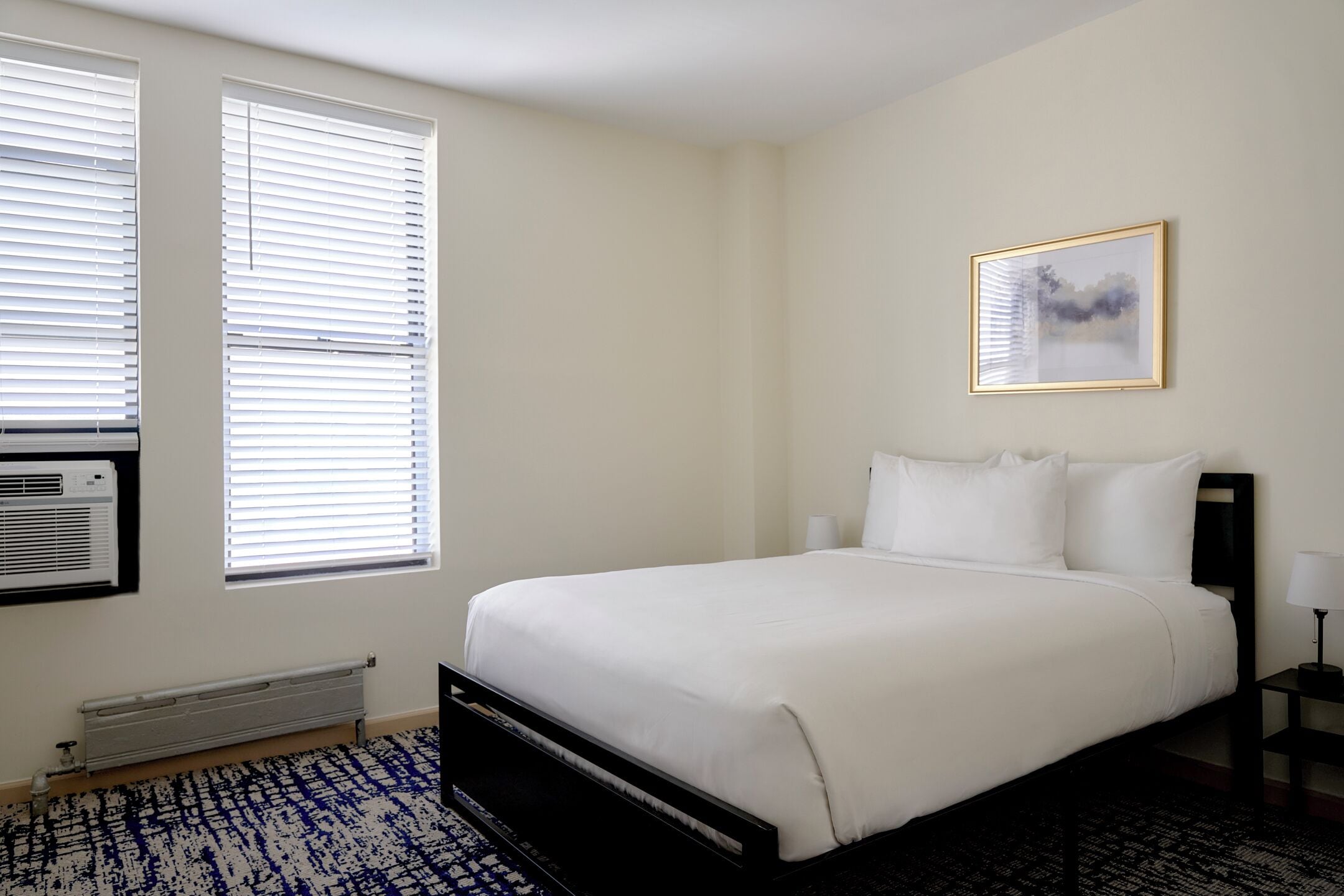 Un dormitorio con cama, dos ventanas con persianas, un radiador, aire acondicionado instalado en la pared, un cuadro enmarcado y una mesita de noche con lámpara.