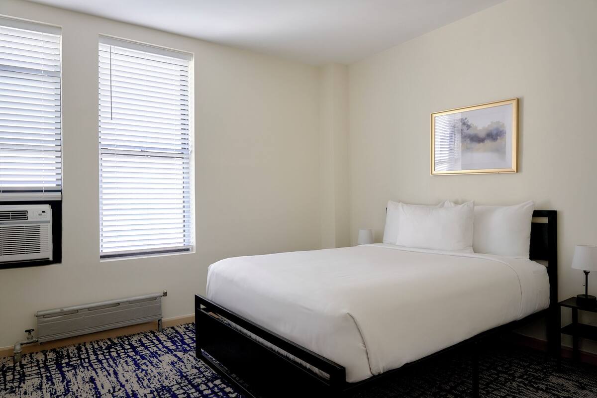 A bedroom with a large bed, two windows with blinds, a radiator, a wall-mounted air conditioner, a framed picture, and a bedside table with a lamp.