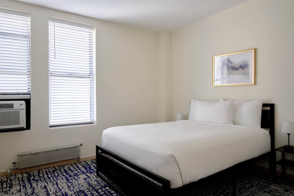 Un dormitorio con cama, dos ventanas con persianas, un radiador, aire acondicionado instalado en la pared, un cuadro enmarcado y una mesita de noche con lámpara.