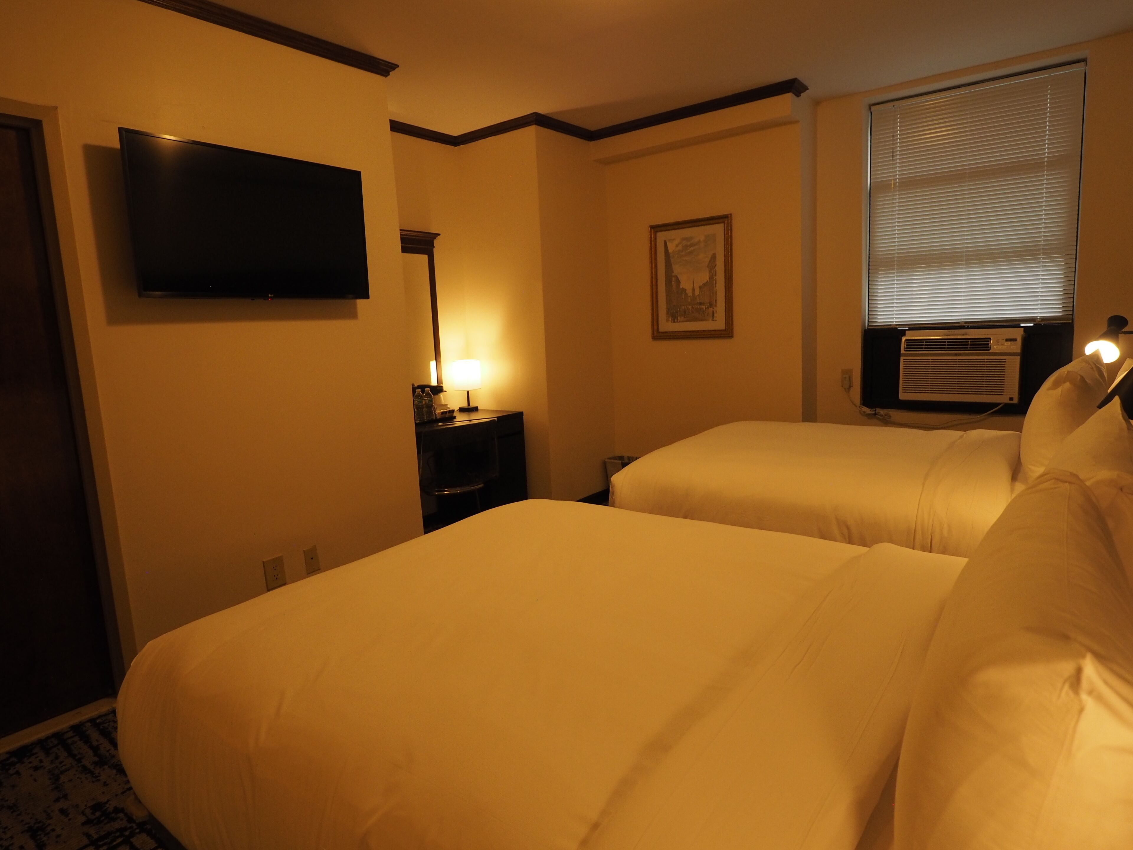 Habitación de hotel con dos camas, un televisor de pantalla plana montado en la pared, una mesita de noche con lámpara, un cuadro enmarcado y un equipo de aire acondicionado.