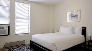 Un dormitorio con una cama grande, dos ventanas con persianas, un radiador, aire acondicionado de pared, un cuadro enmarcado y una mesita de noche con lámpara.