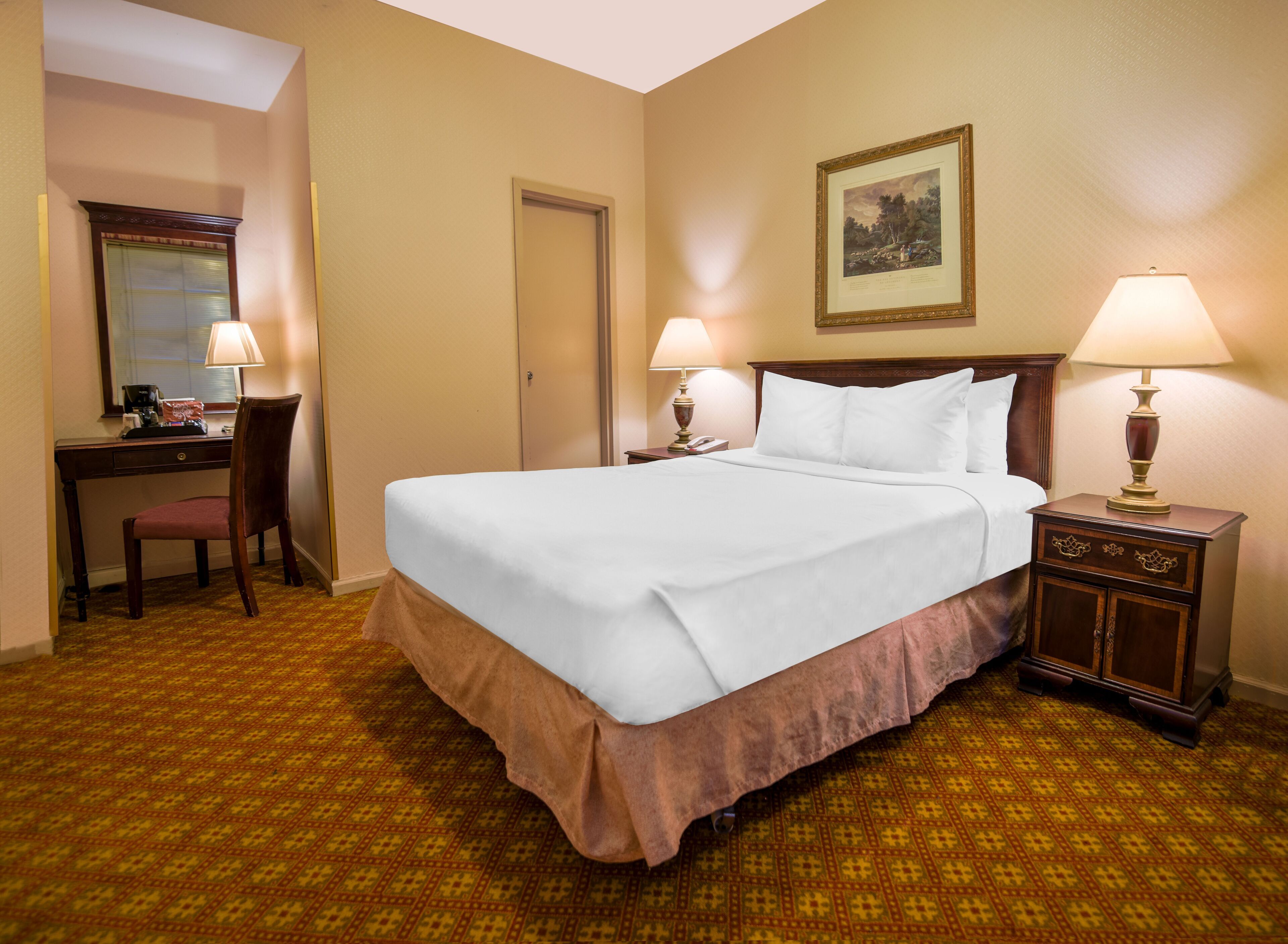 A hotel room with a large bed, two bedside tables with lamps, a desk with a chair, a door, and a framed picture on the wall.