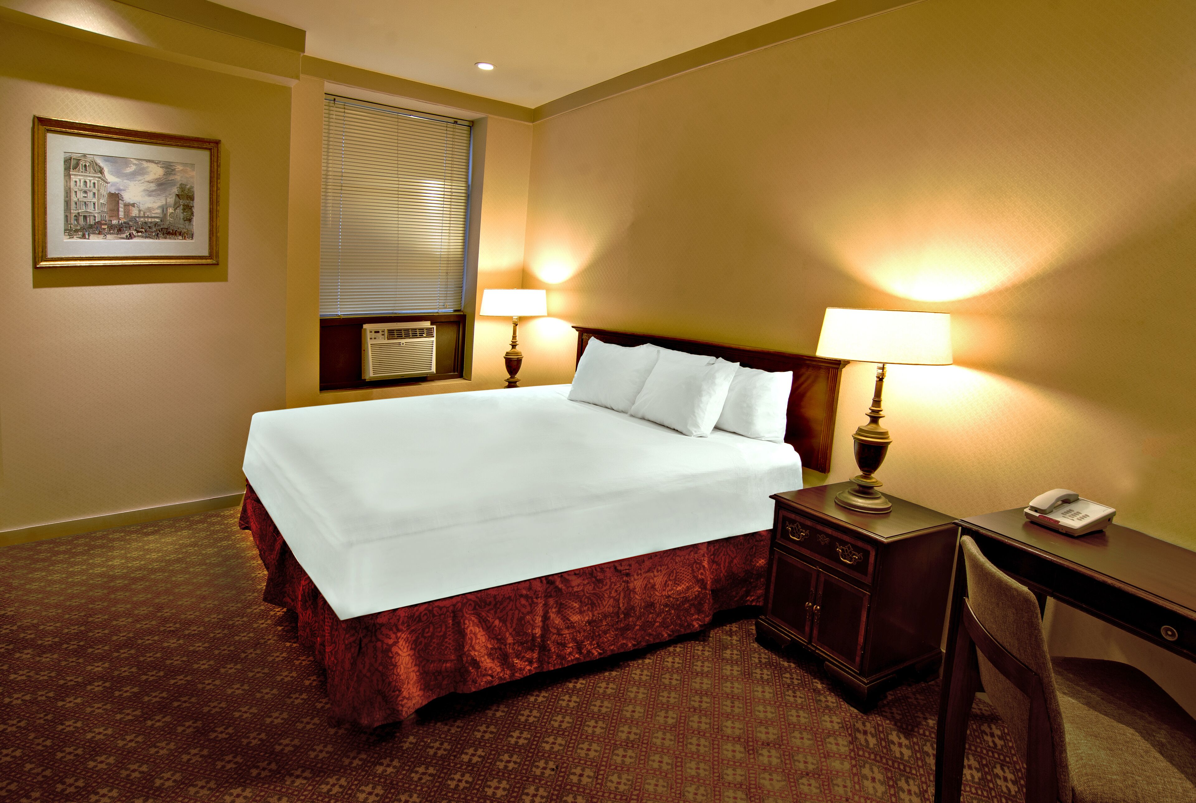 A hotel room with a large bed, two bedside tables with lamps, a desk with a telephone, and a framed picture on the wall.