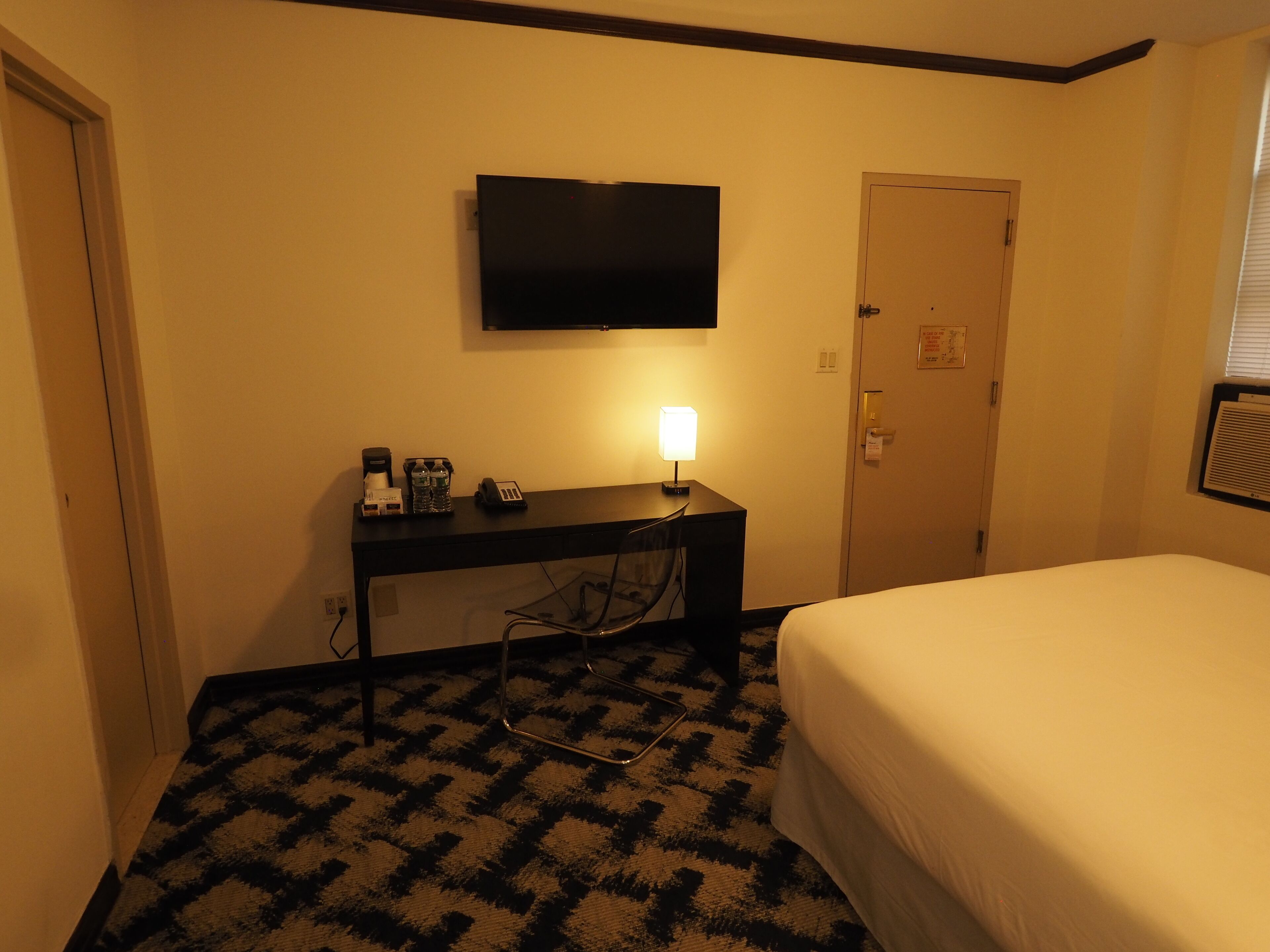A hotel room with a bed, a desk with a lamp, a flat-screen TV, and a door.