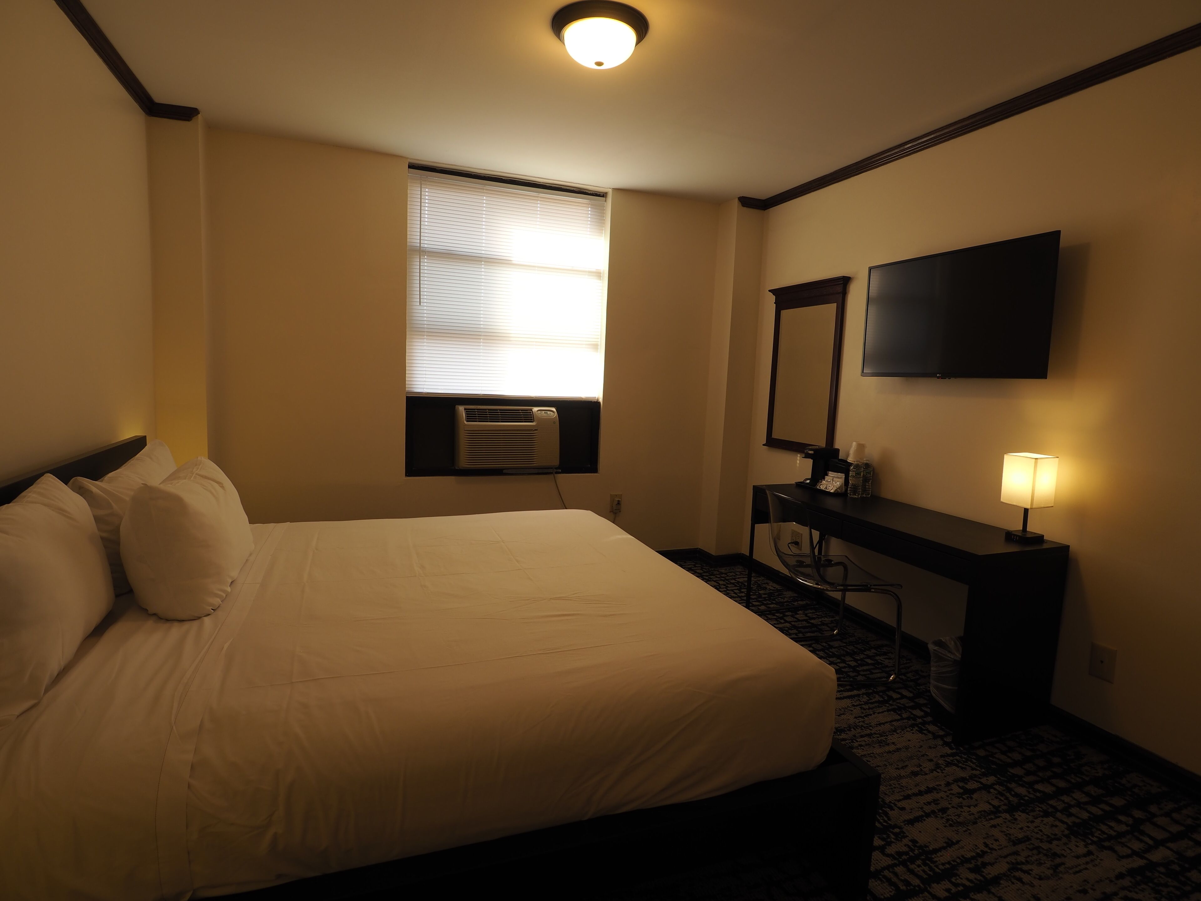 Habitación de hotel con cama, escritorio, silla, televisión de pantalla plana, ventana con persianas y luz de techo.