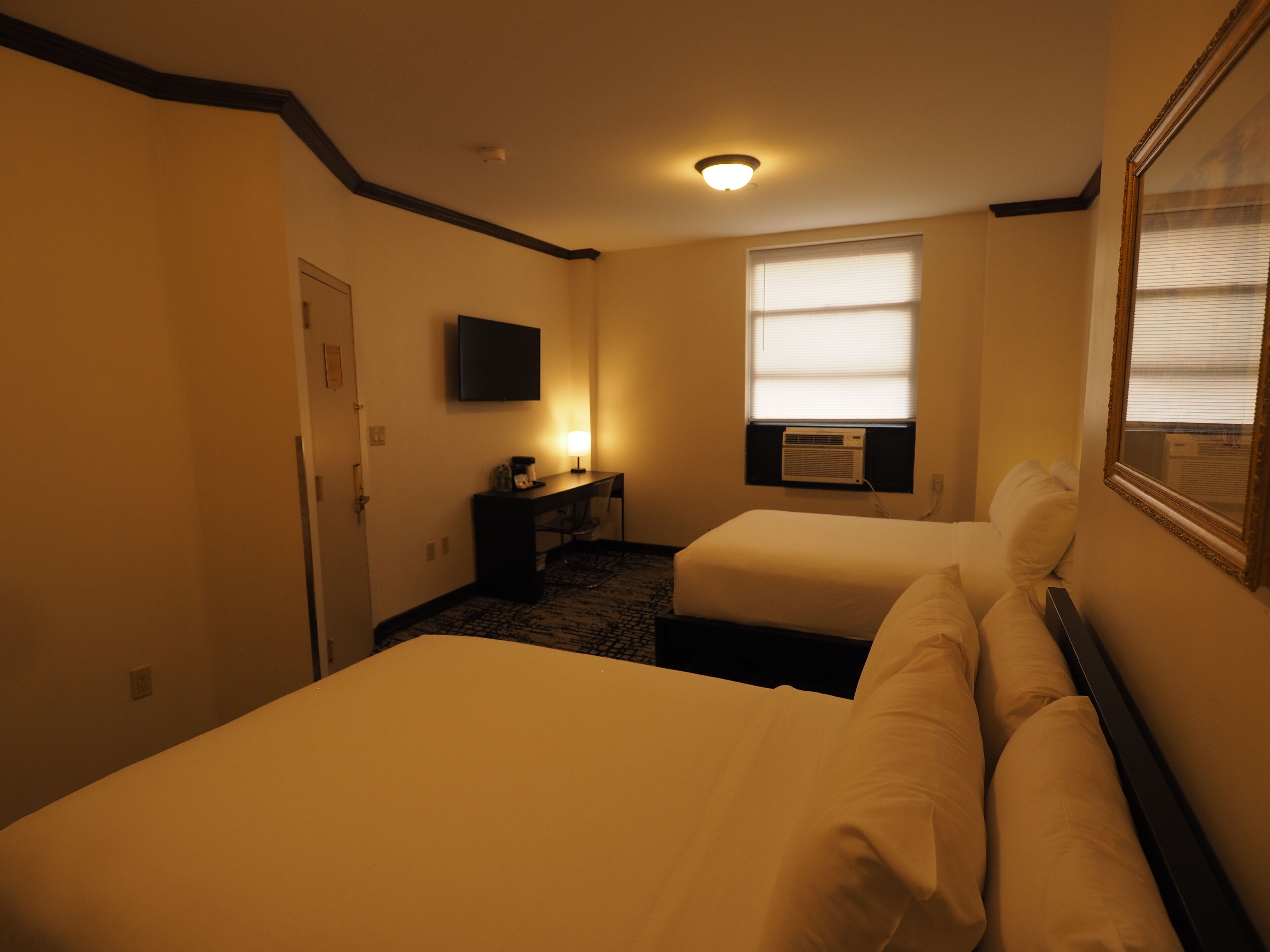 Habitación de hotel con dos camas, un escritorio con lámpara, un televisor en la pared y un equipo de aire acondicionado.