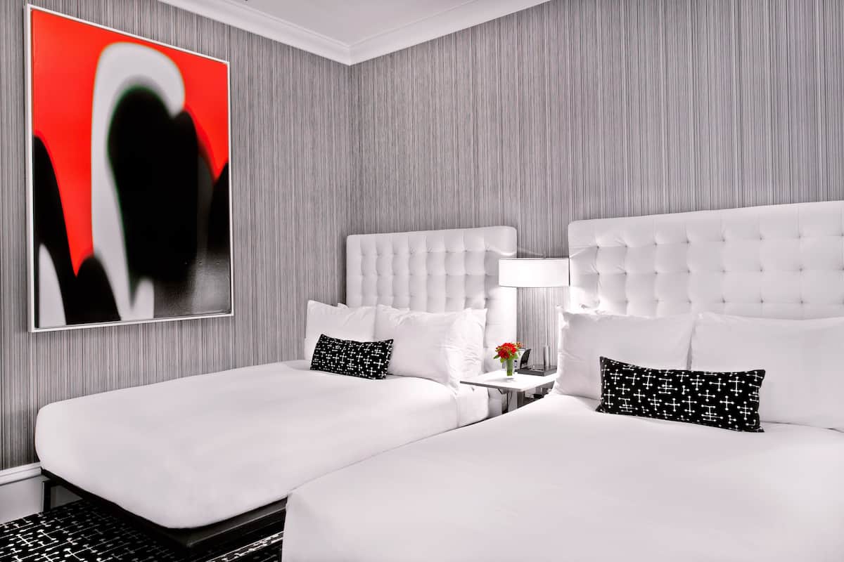 Quarto de hotel com duas camas, uma pintura abstrata grande, uma mesa de cabeceira com abajur e uma pequena mesa com um vaso de flores.