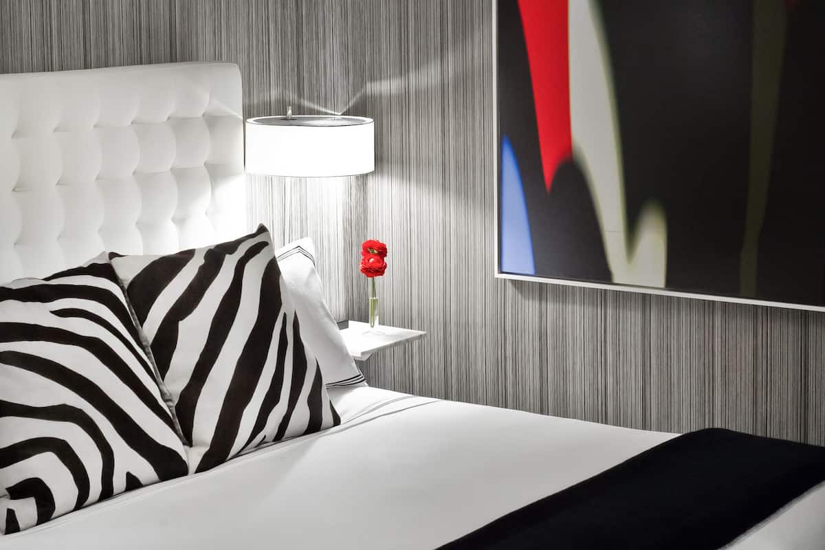 Quarto de hotel moderno com cabeceira estofada, uma grande pintura abstrata, um vaso de flores vermelho e uma almofada listrada.