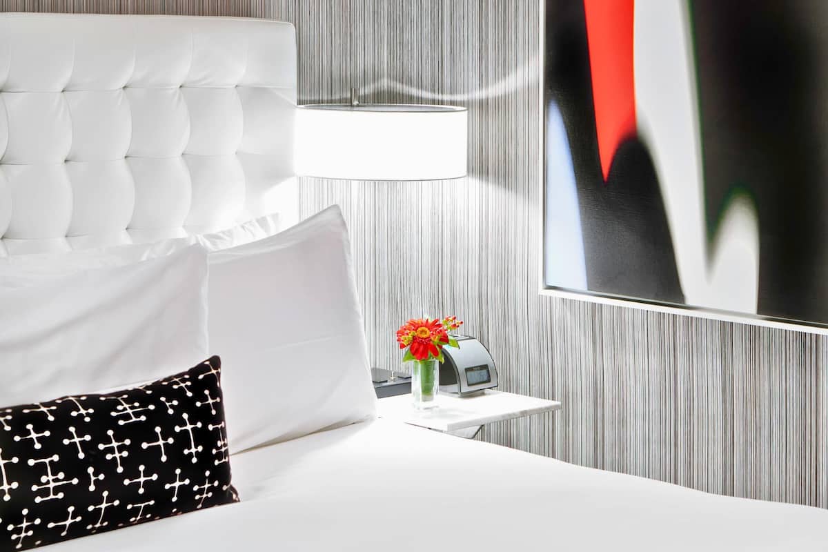 Quarto de hotel com cabeceira estofada, cama branca, travesseiro preto com cruzes brancas, mesa de cabeceira com abajur e quadro na parede.