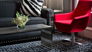 Una habitación de hotel moderna con un sofá oscuro, una silla roja, un tapete con estampado blanco y negro, y un cuadro abstracto grande en la pared.