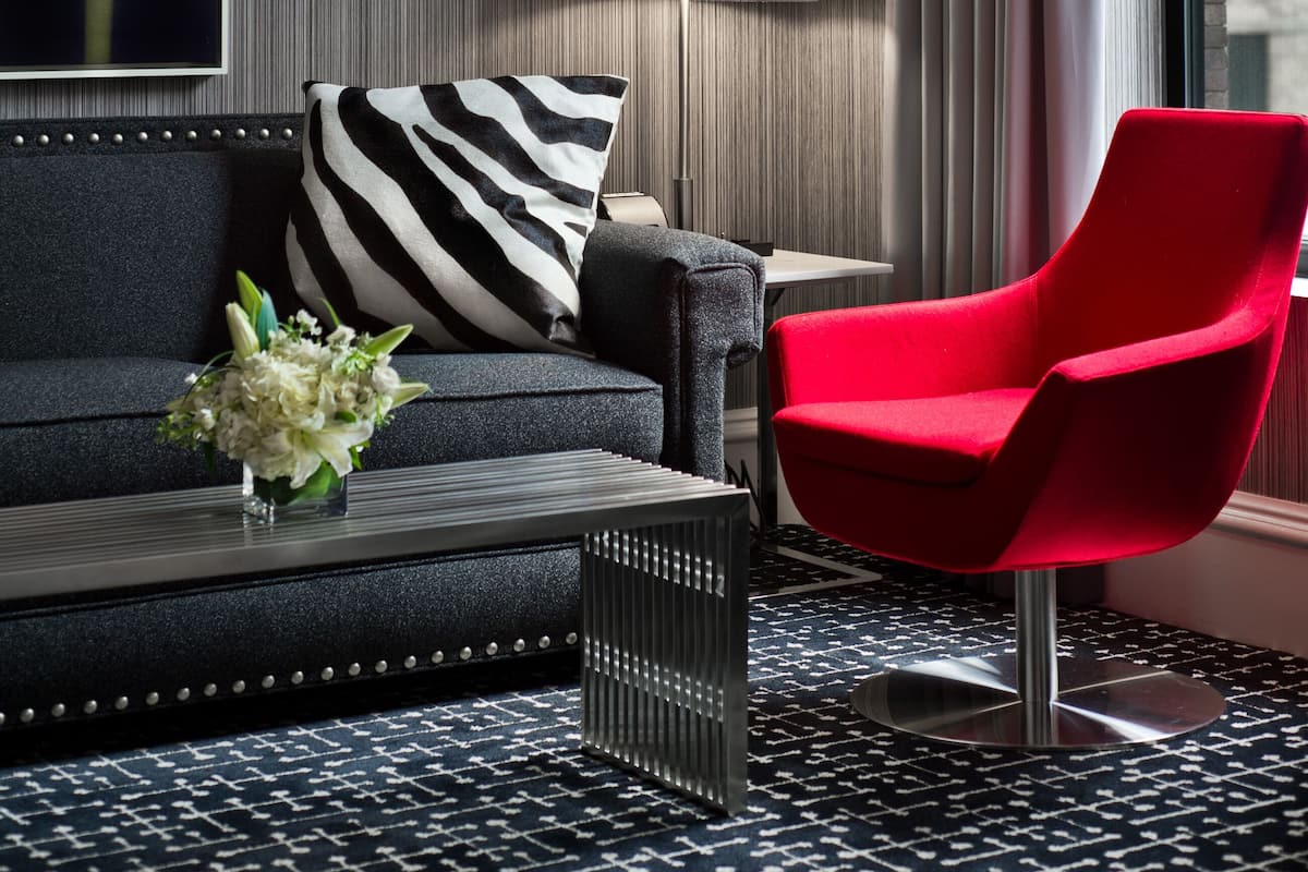 Quarto de hotel moderno com um sofá escuro, uma poltrona vermelha, um tapete com padrão preto e branco e uma grande pintura abstrata na parede.