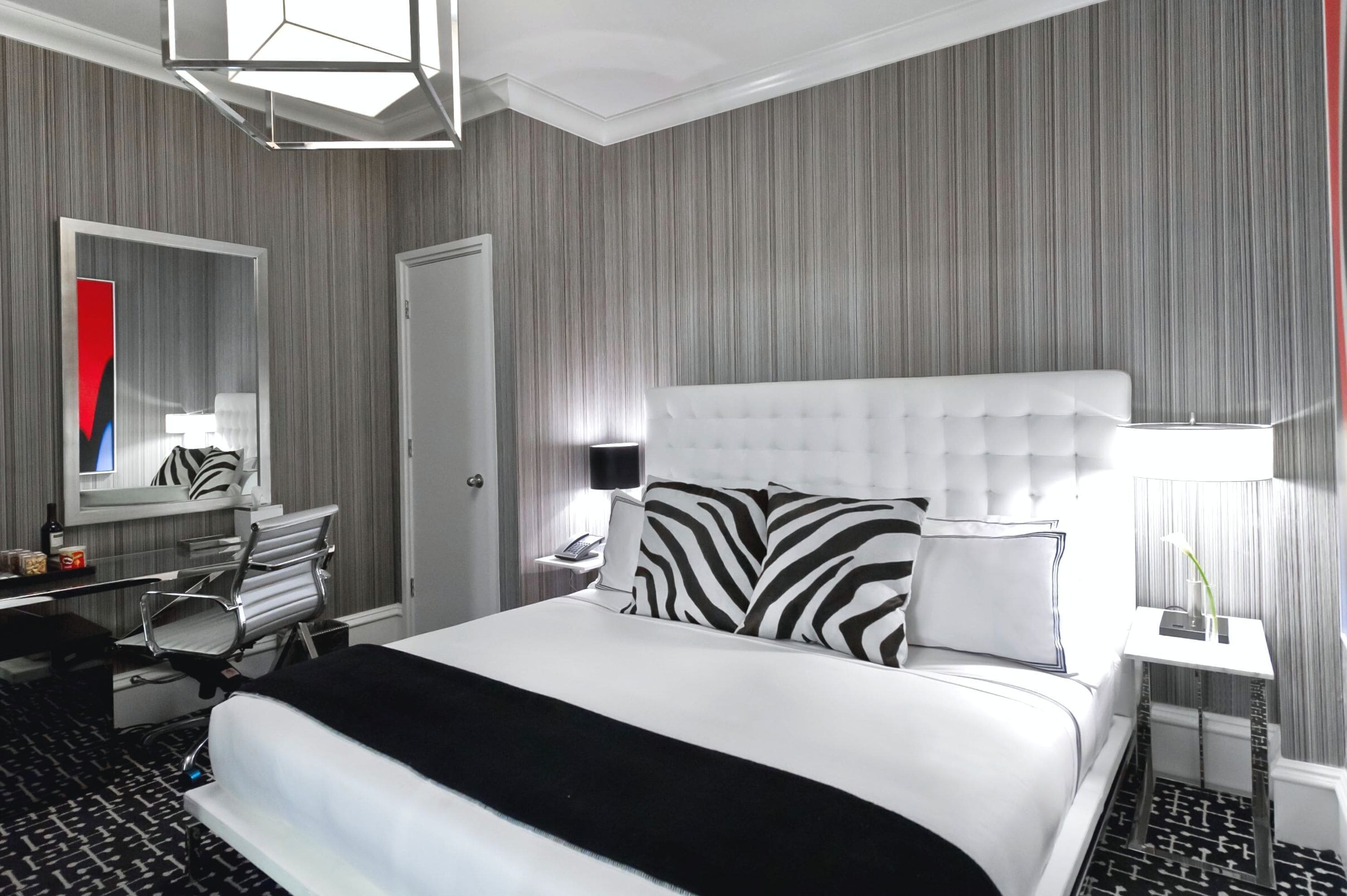 A hotel room with a large bed, a desk, a chair, a mirror, and a nightstand with a lamp.
