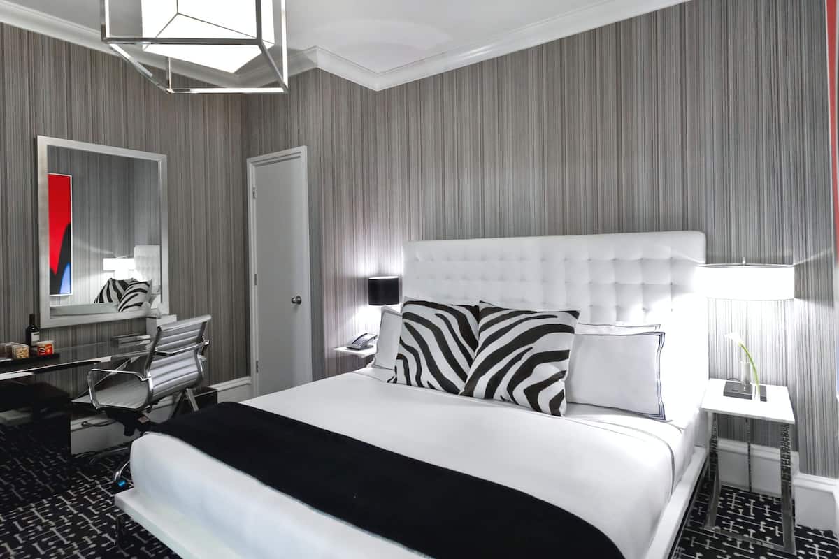 A hotel room with a large bed, a desk, a chair, a mirror, and a nightstand with a lamp.
