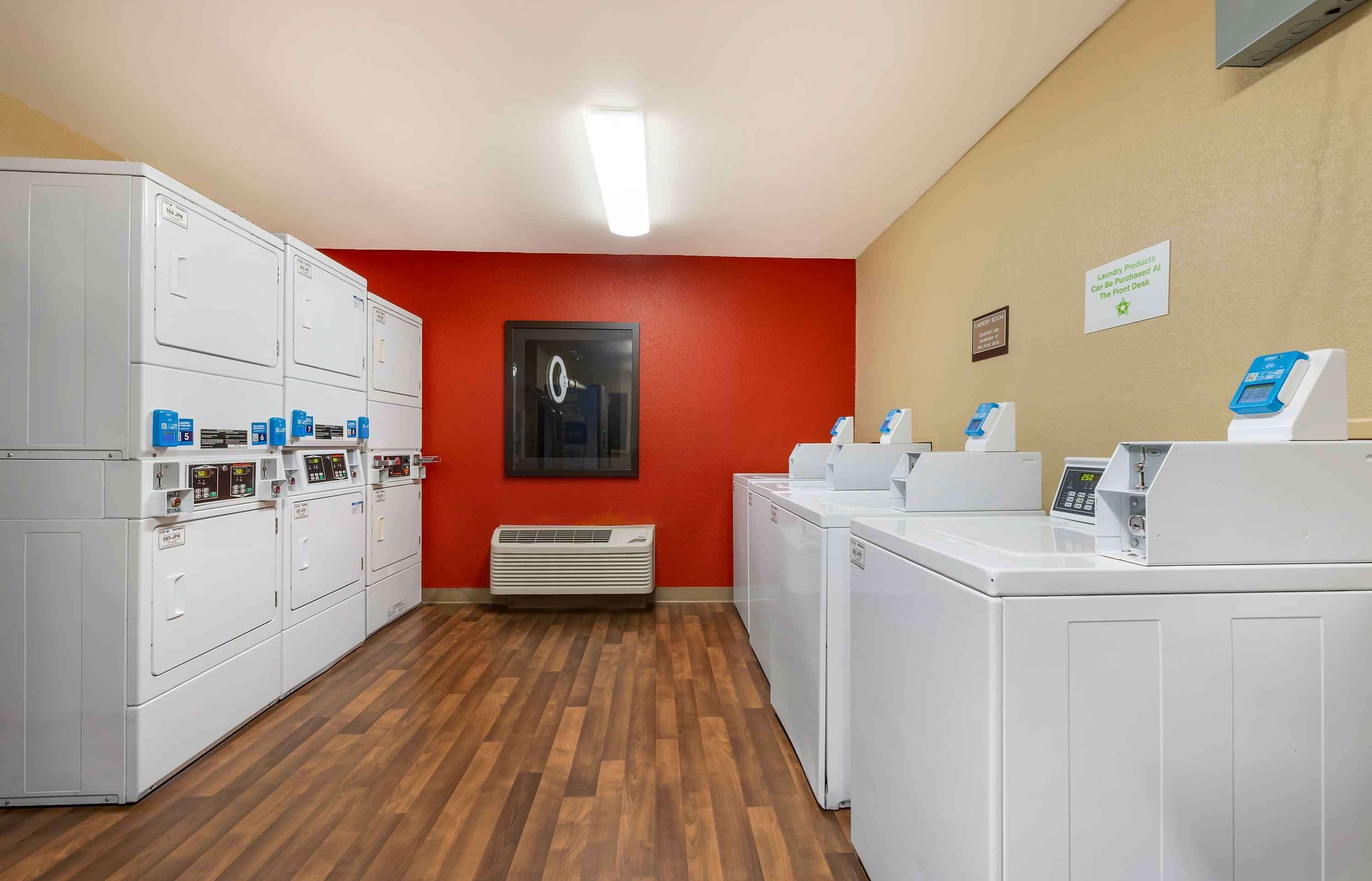 Une buanderie équipée de plusieurs machines à laver et de plusieurs sèche-linge, avec une horloge au mur et un panneau indiquant que les produits de lessive sont en vente à la réception.
