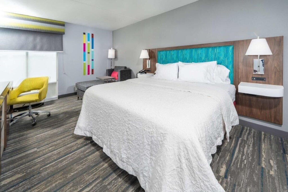 A hotel room with a large bed, a desk with a chair, a small sofa, and a nightstand with a lamp.