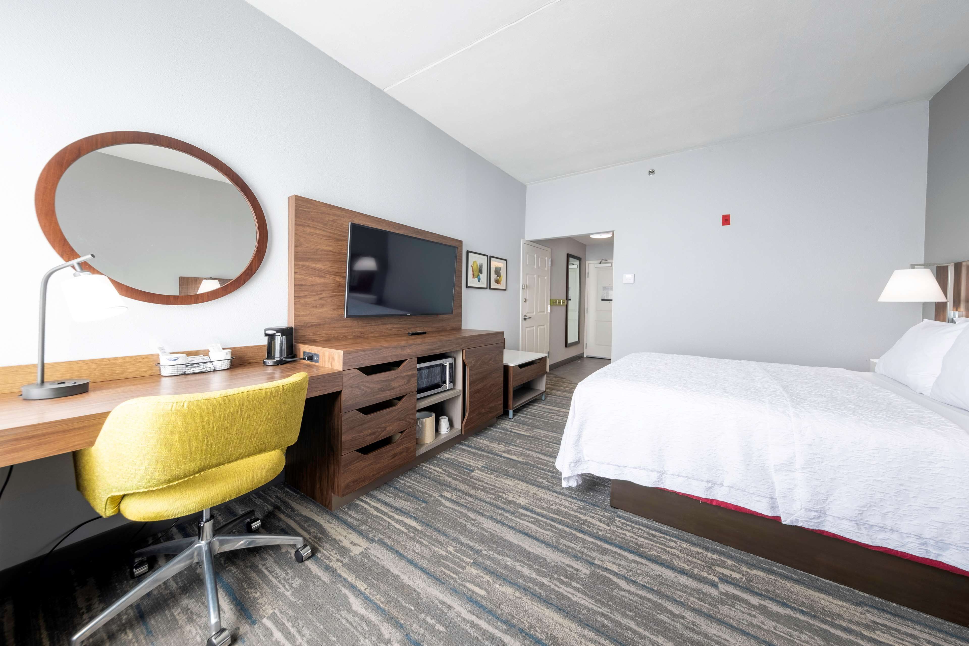 Une chambre d’hôtel comprenant un lit, un bureau avec une chaise, une télévision, un miroir et un sol recouvert de moquette.