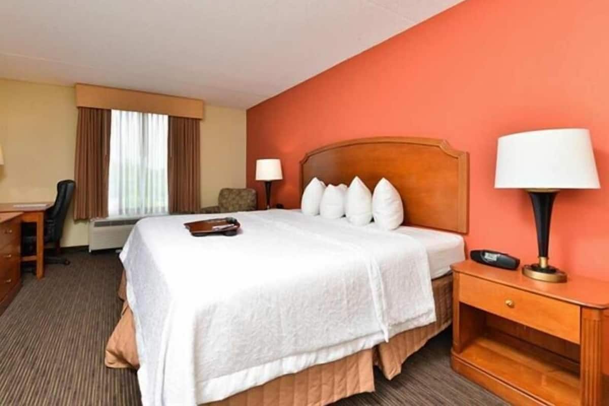 A hotel room with a bed, bedside table, lamp, chair, desk, and window with curtains.