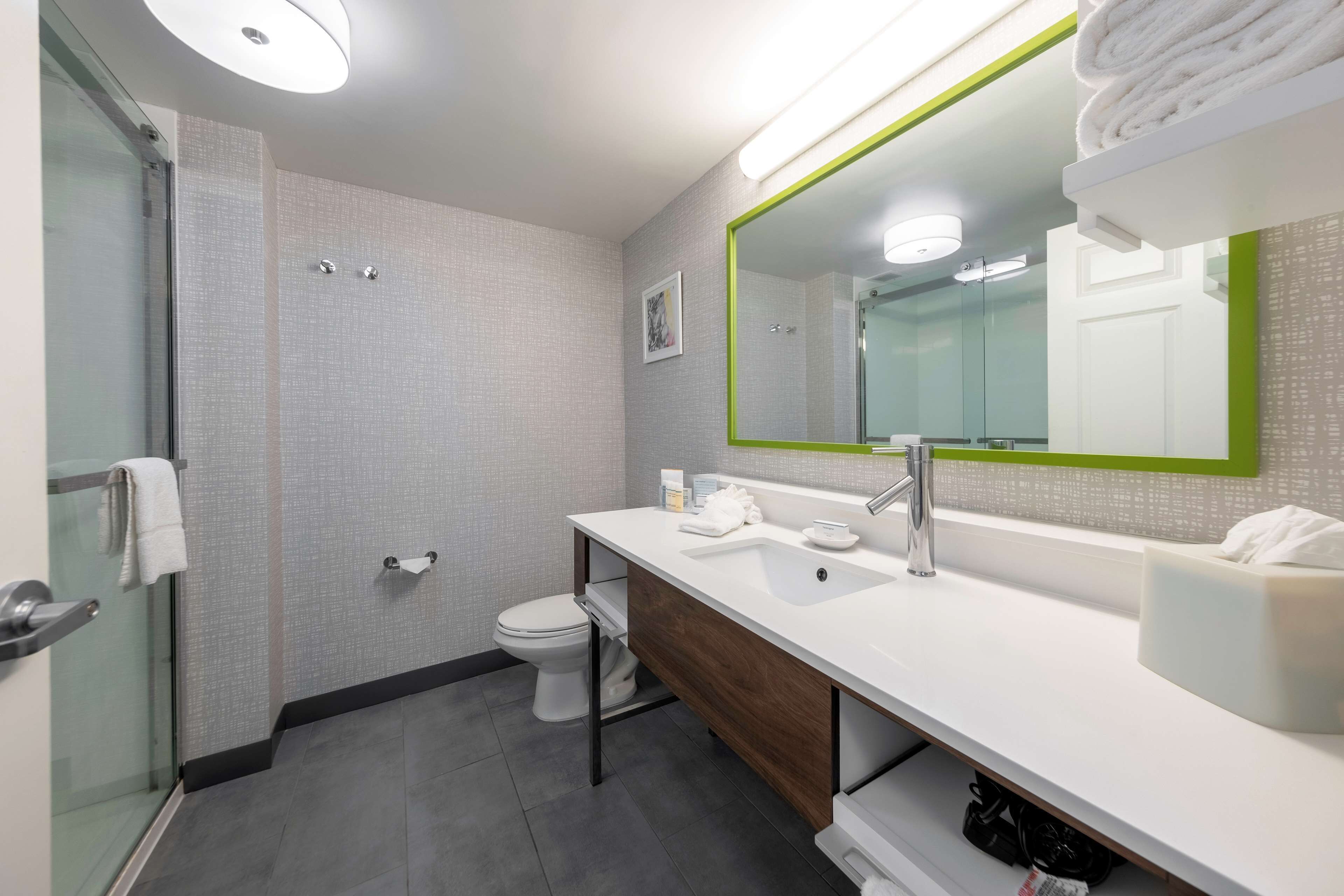 Un baño moderno con cabina de ducha de vidrio, un lavabo blanco con espejo de marco verde y un inodoro.